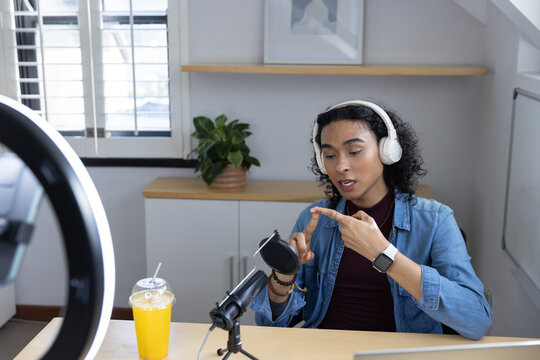 Non-binary person wearing denim jacket and white headphones speaking into condenser mic at desk
