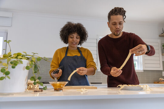 Couple wearing knit sweaters with apron, pastry brush preparing pie lattice at kitchen counter