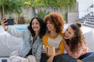 Obraz premium Diverse female friends taking selfie on light sofa by pool, holding smartphone and ceramic mug