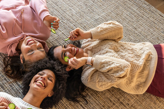 Diverse female friends lying head-to-head on rug wearing hoodie and sweater holding cucumber slices