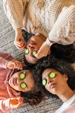 Diverse female friends lying on woven rug at home with cucumber slices, wearing knit sweaters
