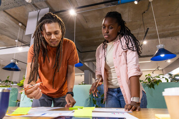 Naklejka premium Diverse coworkers leaning on table checking diagrams pointing at sticky notes with coffee in office