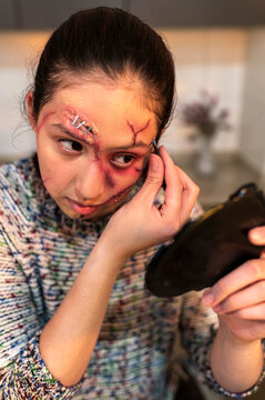 Young woman with realistic gash and staples checking bruised eye makeup in mirror