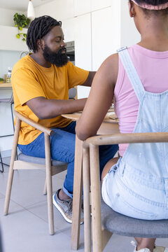 African father and teenage daughter sitting and talking at home kitchen dining table with plates