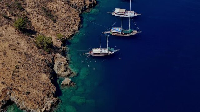 Aerial view of traditional gulet boats anchored near a rocky coastline in clear turquoise water, ideal for summer travel, coastal tourism, sailing, vacation and Mediterranean lifestyle content.