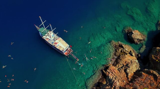 Aerial view of tour boats anchored in clear turquoise water near a rocky coast with people swimming, ideal for summer travel, tourism, vacation, sailing and Mediterranean lifestyle content.