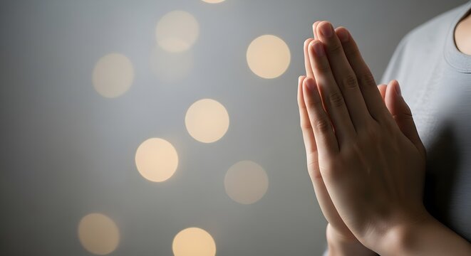 Person performing a prayer gesture with hands clasped together, illuminated by soft, blurred bokeh lights in the background