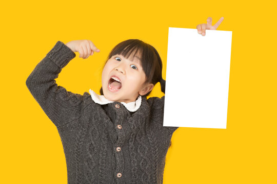 Surprised little girl pointing her finger at a blank white paper sheet with an open mouth expression on a vivid yellow background.