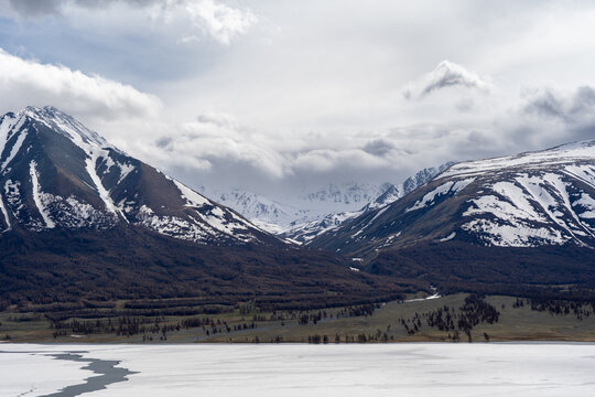 Frozen Alpine Lake with Melting Ice, Larch Forest and Snow-Capped Mountain Range Under Dramatic Clouds, Mongolian Altai