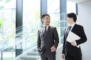 新緑のオフィスの窓際で会話する若手ビジネスマンと上司、取引先のコミュニケーション商談や指導ojtなどの仕事イメージの上半身 © kapinon