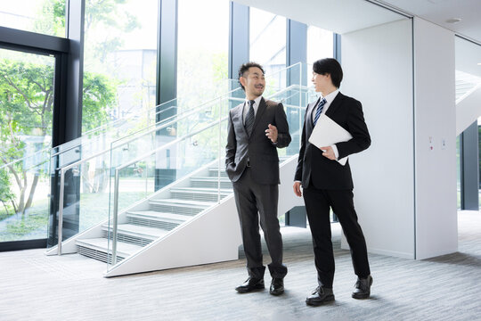 新緑のオフィスの窓際で会話する若手ビジネスマンと上司、取引先のコミュニケーション商談や指導ojtなどの仕事イメージの全身広角