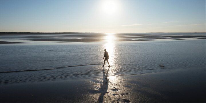 A solitary figure walks along a vast tidal flat at sunset, casting a long shadow on damp sand amidst a serene atmosphere.