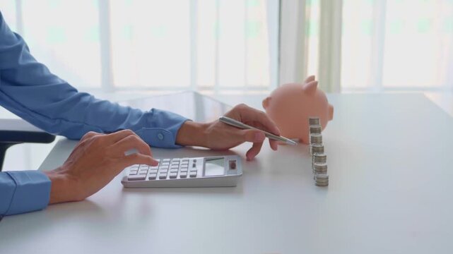 Asian businessman pressing calculator and holding pen counting stacking coin with piggy bank.