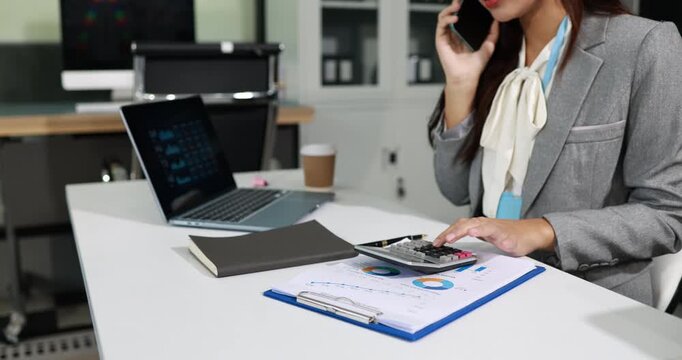 Businesswoman in a gray suit talking on the phone while working at an office desk with calculator, laptop, notebook and financial charts