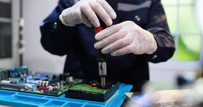 Technician in gloves repairing a hard drive with a screwdriver on a workbench, with circuit boards in the background in a clean electronics lab.