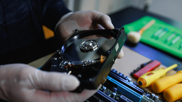 Close-up of gloved hands holding an open hard drive above a computer motherboard with repair tools, showing data recovery, hardware service, and technology maintenance.