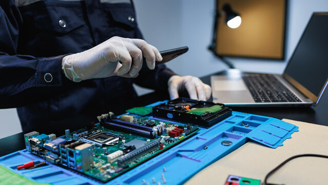 Technician in gloves photographing computer parts on a workbench with motherboard, hard drive and laptop, showing electronics repair, data recovery and hardware diagnostics.
