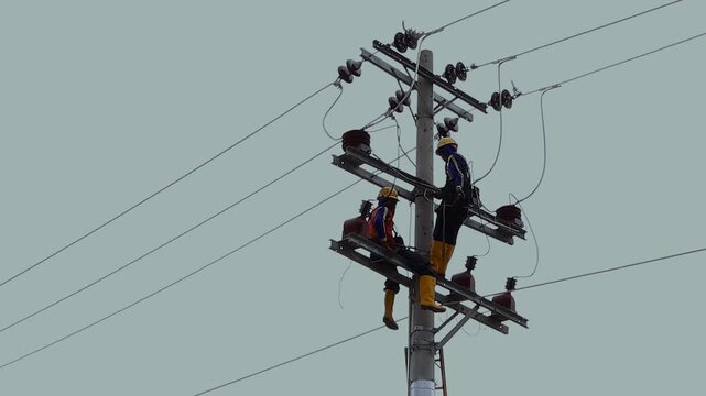 Two electricians work on a tall utility pole. They wear helmets and safety gear. One stands, adjusting wires. The other sits, handling equipment. They focus on their task. Use for construction