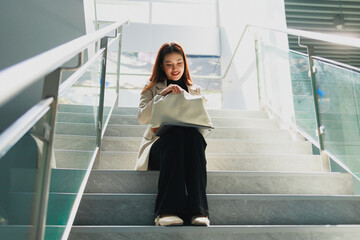 Obraz premium Young woman sitting on indoor stairs in a bright modern building, looking into her handbag. Casual fashion, everyday lifestyle, calm mood, natural light, and clean office interior.