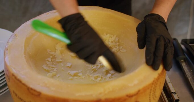 Gourmet cooking. Cheese food. Chef hand in black gloves scraping parmesan wheel inside with metal spoon preparing dish ingredient.