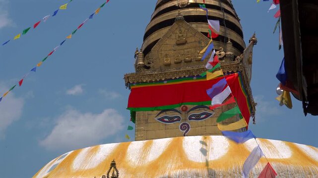 A static low-angle medium shot of the golden spire and wisdom eyes of Swayambhunath Stupa with colorful Buddhist prayer flags fluttering actively in Kathmandu, Nepal