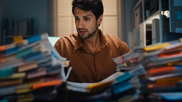 Overworked young Latin businessman working late on document in large stack of paperwork files at office night. Tax audit deadline, accounting stress, corporate burnout, urgent business concept.