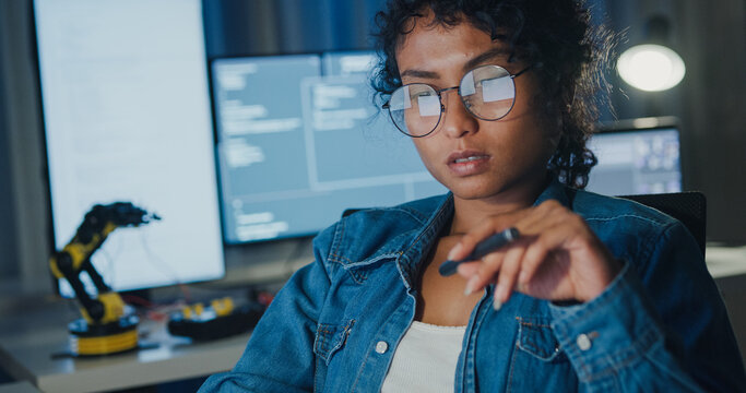 Close-up young Black female programmer coding AI software on multiple screens in night office. Network security, cybersecurity, machine learning, working late, overtime, robotics automation concept.