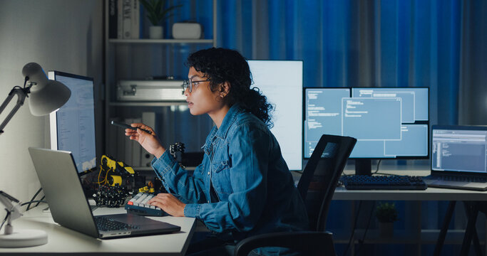 Young African American female programmer coding AI software on multiple screens in night office. Network security, cybersecurity, machine learning, working late, overtime, robotics automation concept.