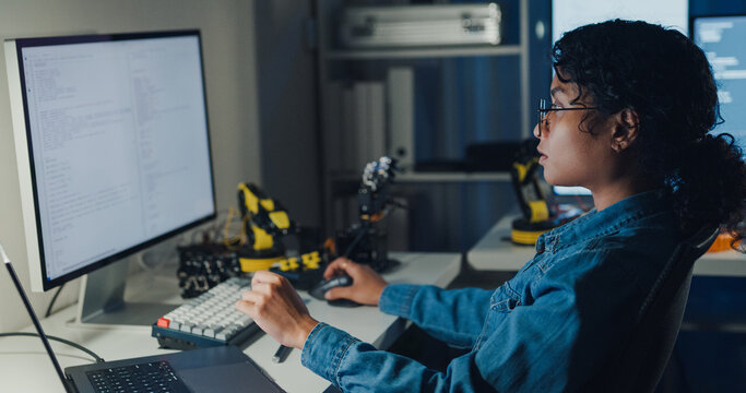 Young African American female programmer coding AI software on multiple screens in night office. Network security, cybersecurity, machine learning, working late, overtime, robotics automation concept.