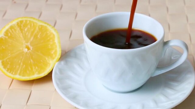 Pouring hot black coffee or espresso into a white textured cup next to a fresh lemon slice.
