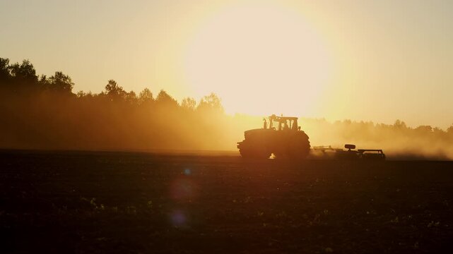 Silhouette of agricultural tractor cultivating farmland at sunset. Modern farming machinery preparing soil and sowing crops in rural countryside under golden evening light.