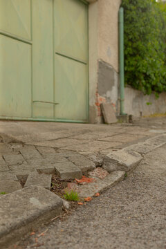 Low-angle view of a cracked concrete driveway edge with loose paving slabs and small weeds growing between them, leading to a pale green metal garage door set