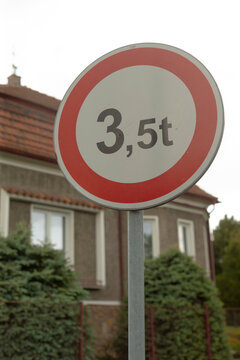Circular traffic sign with a red border showing a 3.5t weight limit mounted on a metal post in front of a suburban house. Foreground sign is in sharp focus