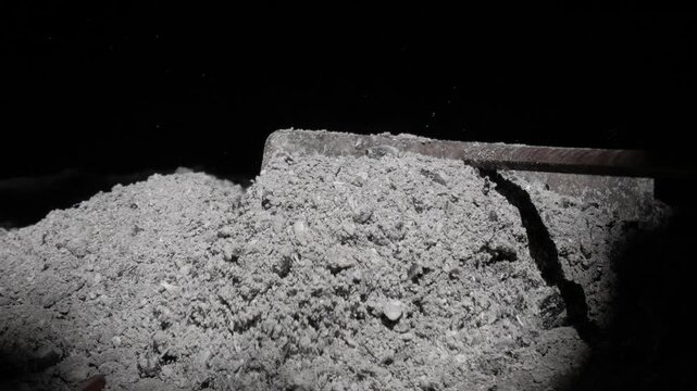Metal tool rakes pile of ash in dimly lit indoor setting, showing fine debris texture