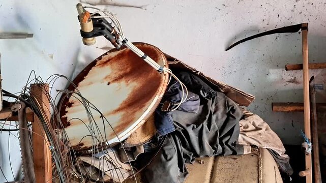 Wide view of an old rusty satellite dish among scattered junk and discarded items inside a cluttered garage or workshop shed. Rustic atmosphere, neglect, storage, decay, and abandoned equipment