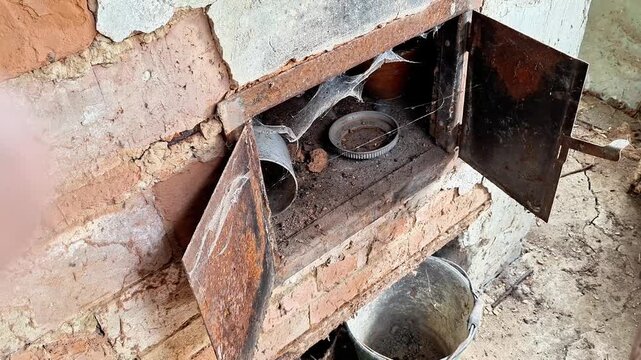 Old rustic brick stove with open metal doors in an abandoned building. Dust, debris, and signs of decay create an authentic atmosphere of neglect, age, and rural abandonment