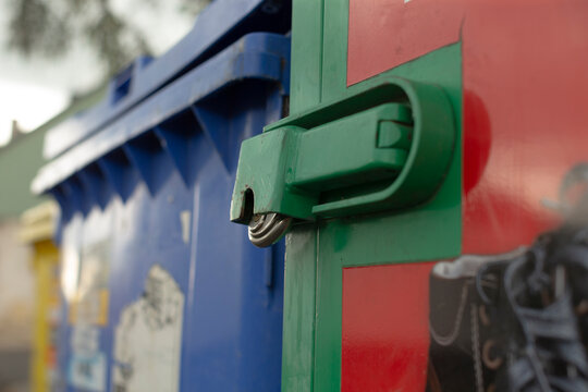 Shallow depth-of-field close-up showing a green metal latch secured on the side of a red recycling container. A blue plastic waste bin and other out-of-focus