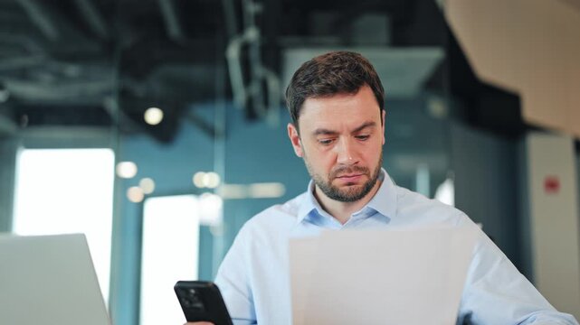 Close-up of happy businessman or IT specialist checking document and typing on smartphone. Friendly corporate worker, manager or freelance designer smiling at camera at contemporary office desk.
