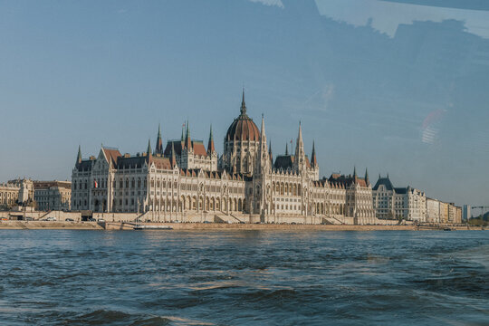 Panoramic view of an ornate historic parliament building with a monumental dome, Gothic Revival towers and detailed facade beside a wide river on a sunny day