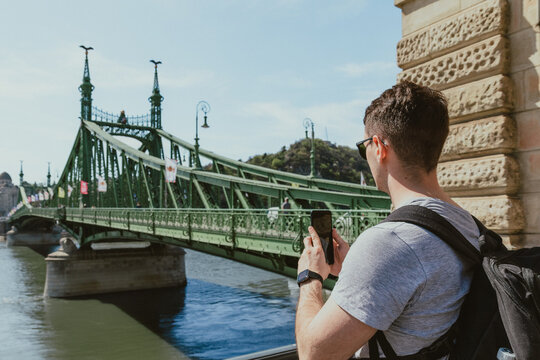 Young male tourist wearing sunglasses and a backpack takes a smartphone photo of the green Liberty Bridge over the Danube River