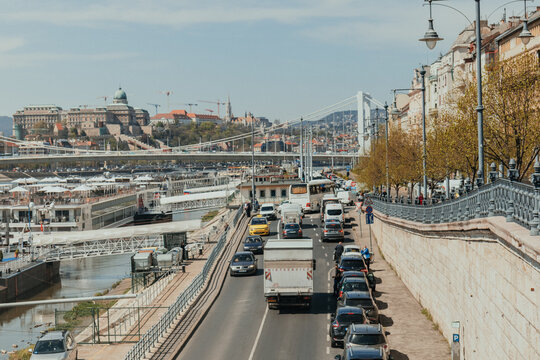 Busy riverside road beside docked cruise ships with suspension bridge and historic city skyline