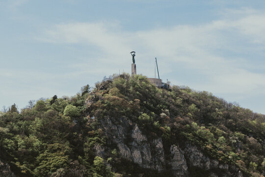 Distant view of the Liberty Statue monument standing on top of a rocky, tree-covered hill, surrounded by spring foliage beneath a clear blue sky