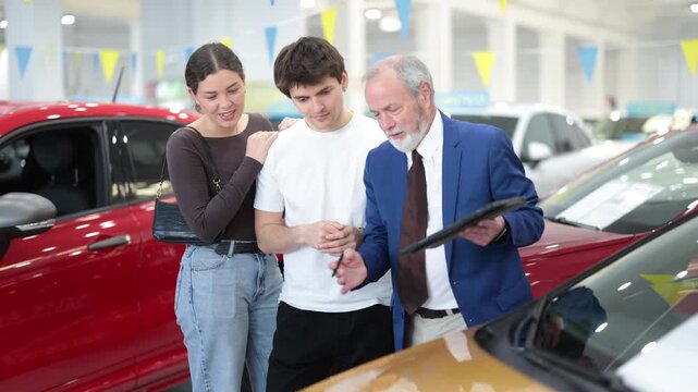 Senior salesman advises a young at a car dealership, showing the car and explaining the details before buying. Buyers choose a machine with the right features and consult with seller. High quality 4k