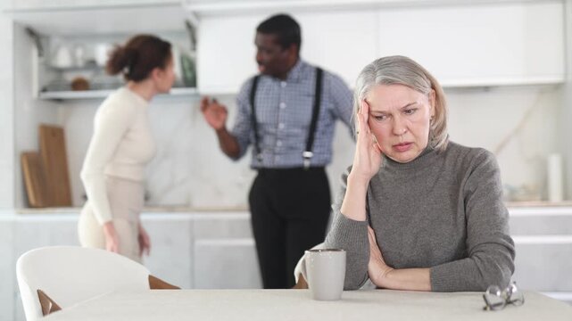Angry mature woman sitting at table while two male and female family members disputing with each other in the back