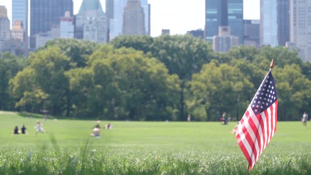 American flag on green grass in Central Park, New York, USA. Sheep Meadow, high-rise buildings and people. Skyline cityscape, Manhattan Midtown skyscrapers. United States symbol and business district.