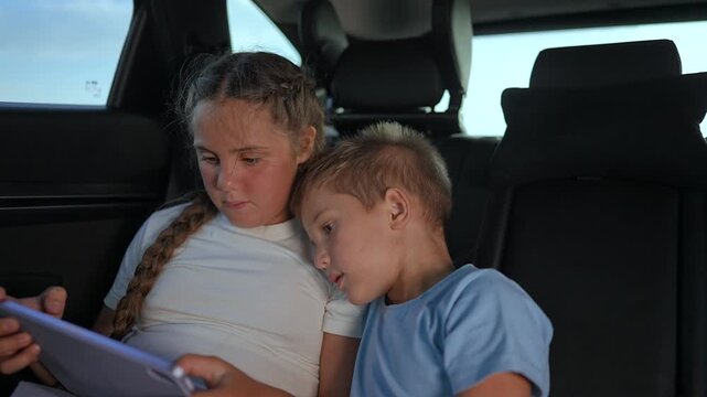 Children watching digital tablet in car. Boy and girl sitting close. Children focus on screen together. Tablet screen keeps children engaged. Smiling children enjoy watching technology in car