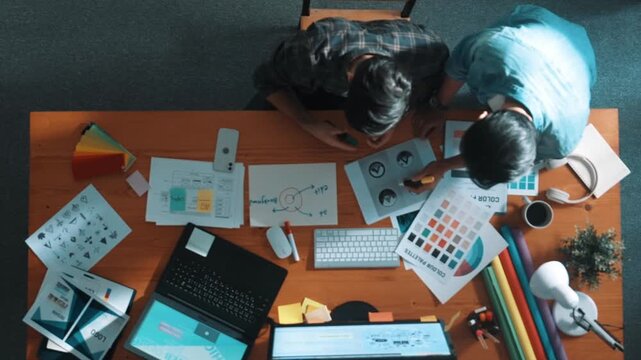 Top down view of smart designer helping manager choosing color from colored palettes. Aerial view of businessman working together and planning together while looking at logo from laptop. Symposium.