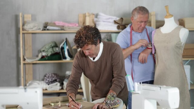 Adult man tailor drawing pattern on paper elderly man assistant sews on mannequin in workshop