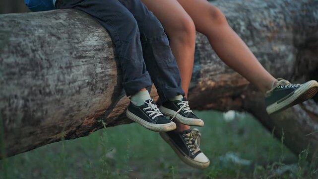 Legs swing gently above forest grass. Boy kicks feet wearing sneakers. Children laugh sitting on log. Feet tap on shoes above forest. Sneakers dangle with children. Sitting legs in forest sneakers.
