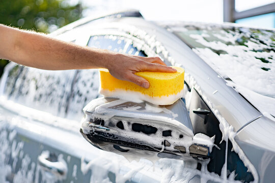 A person's caucasian hand is using a yellow sponge to wash a car's side mirror covered in soap suds